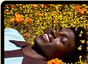 MacBook Neo amarelo-cítrico com a tela mostrando uma pessoa sorrindo deitada em um campo de flores ensolarado