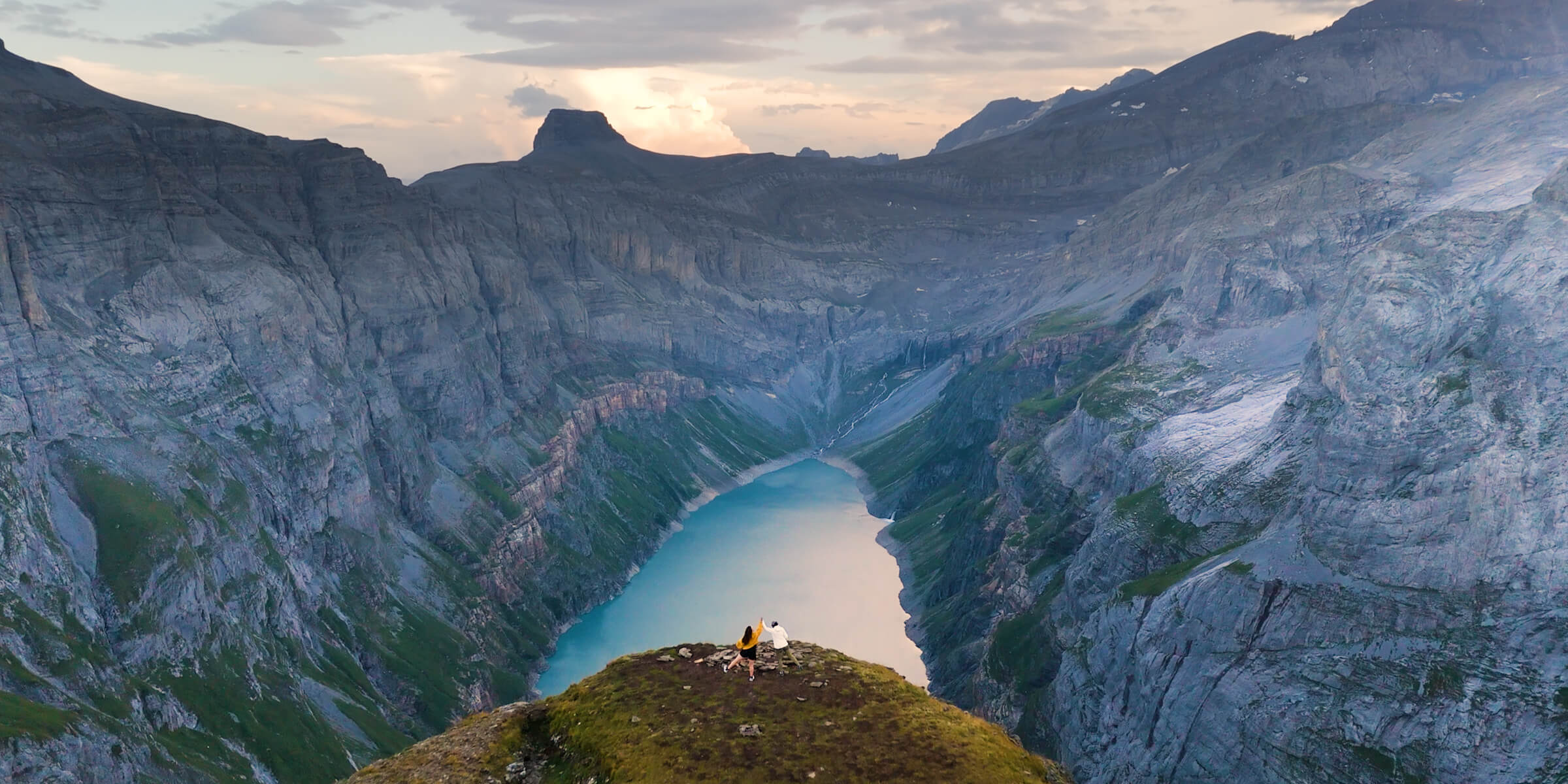 Imagem de um casal no topo de uma montanha e logo em baixo um enorme vale com um grande lago